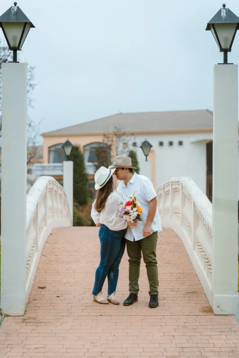 Hombre y mujer permanecen juntos en un puente blanco; la mujer lleva sombrero y sostiene un ramo.