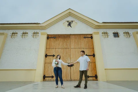 Hombre y mujer permanecen tomados de la mano frente a un gran portón de madera.