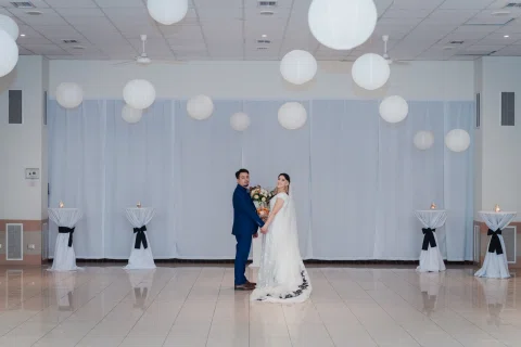 Hombre con traje azul y mujer con vestido blanco posan a distancia en el centro de un salón decorado con globos blancos.