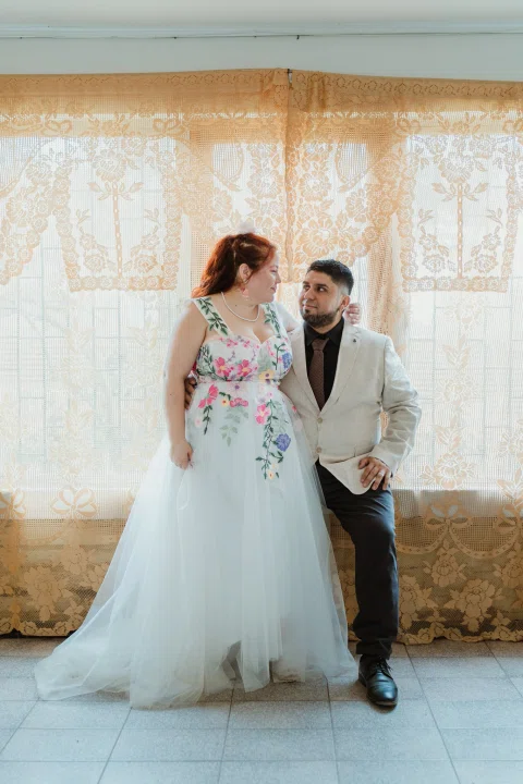 Mujer de cabello rojo con vestido floral y hombre con chaqueta clara posan juntos frente a una cortina clara.