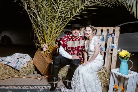 Hombre con poncho y sombrero y mujer con vestido blanco posan sentados junto a una decoración de madera, fardos, flores y hojas de palma.