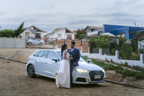 Hombre con traje azul y mujer con vestido blanco posan junto a un automóvil blanco.