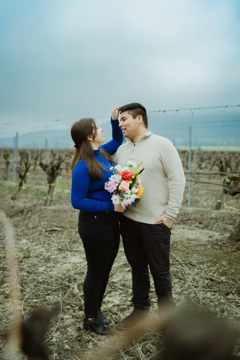 Hombre y mujer posan juntos con un ramo de flores entre hileras de viñas.