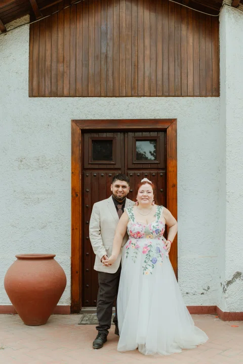 Mujer de cabello rojo con vestido floral y hombre con chaqueta clara posan frente a una puerta de madera.