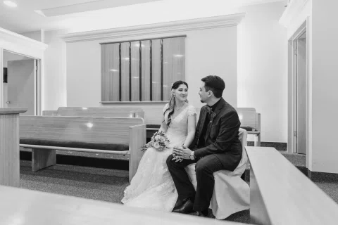 Mujer con vestido blanco y hombre con traje oscuro posan sentados juntos en una banca, en blanco y negro.