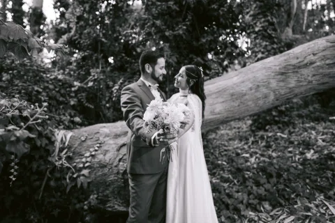 Hombre con traje oscuro y mujer con vestido blanco posan juntos junto a un tronco caído, en una fotografía en blanco y negro.
