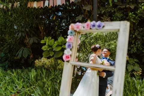 Mujer con vestido blanco y hombre con traje azul se reflejan en un espejo de marco blanco decorado con flores y rodeado de vegetación.