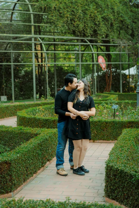 Hombre y mujer se abrazan en un jardín de setos geométricos.
