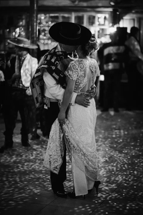 Hombre con poncho y sombrero y mujer con vestido blanco se besan dentro de un salón, en una fotografía en blanco y negro.