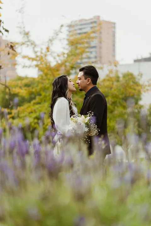 Hombre y mujer se besan detrás de flores violetas desenfocadas en primer plano.