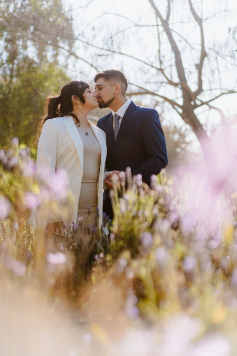 Hombre con traje azul y mujer con traje claro se besan entre flores desenfocadas al aire libre.