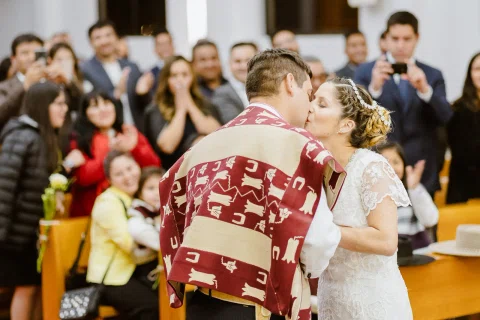 Hombre con poncho y mujer con vestido blanco se besan dentro de una iglesia, con varias personas detrás.