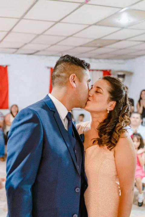 Hombre y mujer se besan en un espacio interior, con varias personas al fondo.