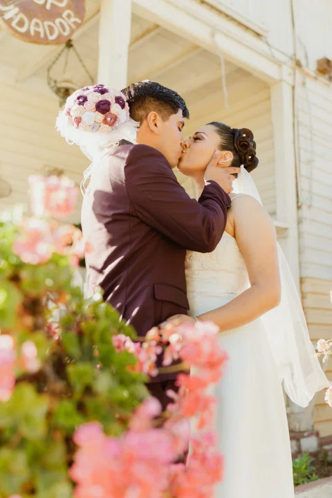 Hombre y mujer se besan junto a flores rosadas en un retrato vertical.