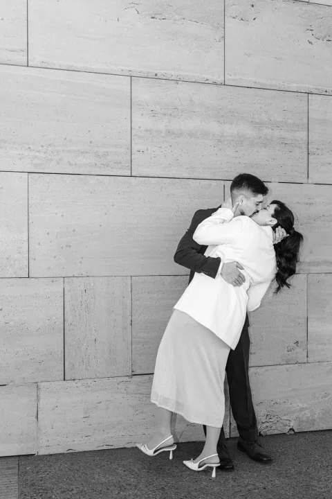 Hombre con traje oscuro y mujer con traje claro se besan frente a un muro, en una fotografía en blanco y negro.