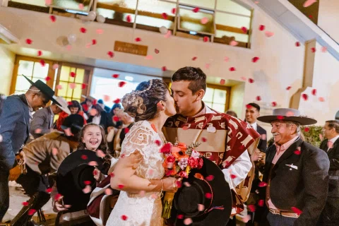 Hombre con poncho y mujer con vestido blanco se besan entre pétalos rojos dentro de una iglesia, rodeados por otras personas.