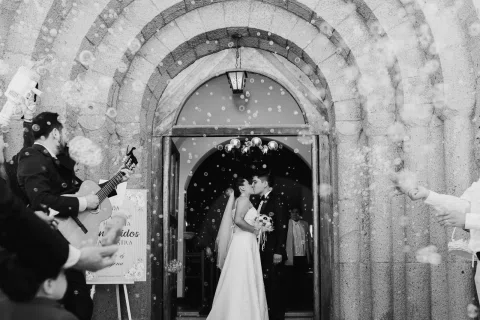 Hombre y mujer se besan en la puerta de una iglesia rodeados de burbujas, en blanco y negro.
