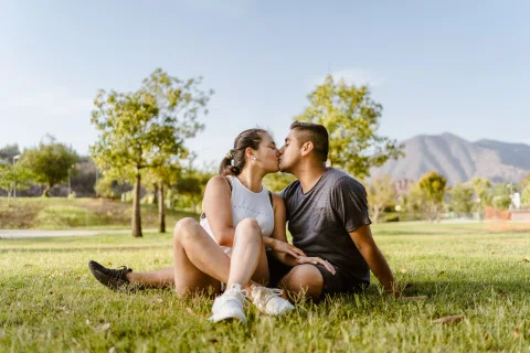 Hombre y mujer se besan sentados en el parque con árboles y montañas detrás.