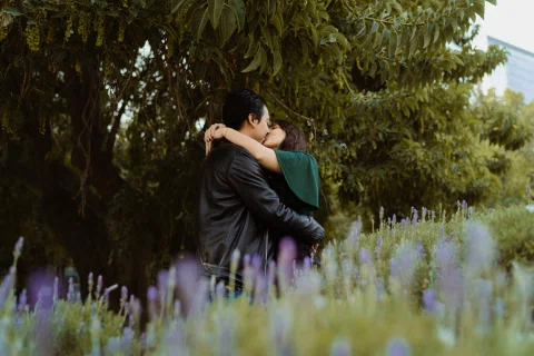 Hombre y mujer se besan detrás de flores lilas desenfocadas en un parque.