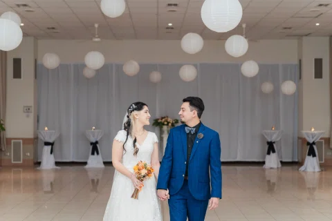 Hombre con traje azul y mujer con vestido blanco se miran mientras posan dentro de un salón.