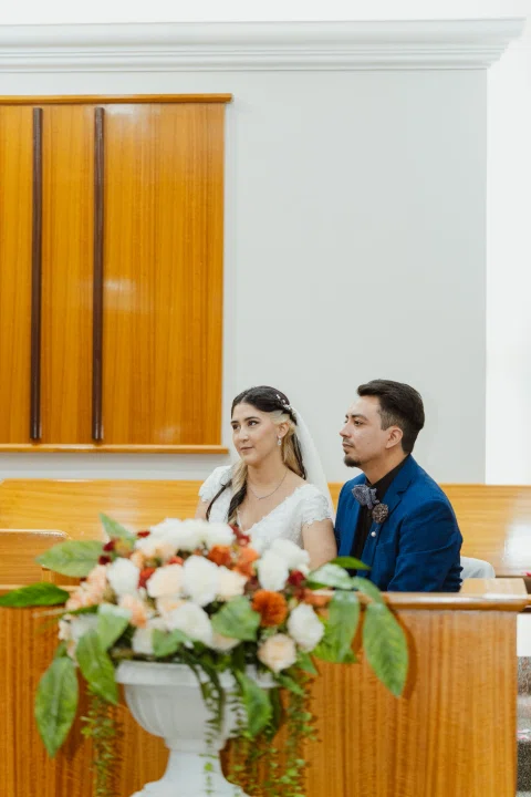 Mujer con vestido blanco y hombre con traje azul permanecen sentados detrás de un arreglo floral en primer plano.
