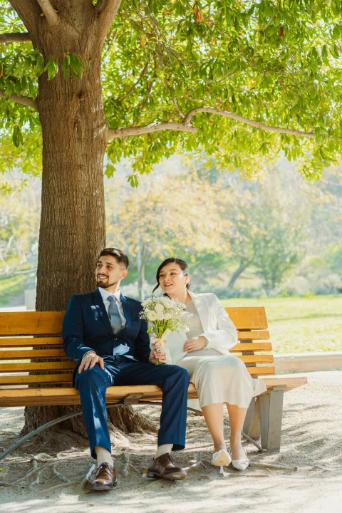Hombre con traje azul y mujer con traje claro están sentados juntos en una banca bajo un árbol; ella sostiene un ramo.