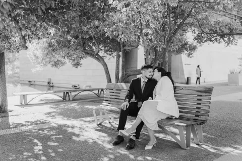Hombre con traje oscuro y mujer con traje claro están sentados juntos en una banca, en una fotografía en blanco y negro.