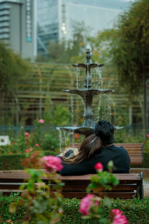 Hombre y mujer están sentados juntos en una banca rodeada de flores, con una fuente cercana.
