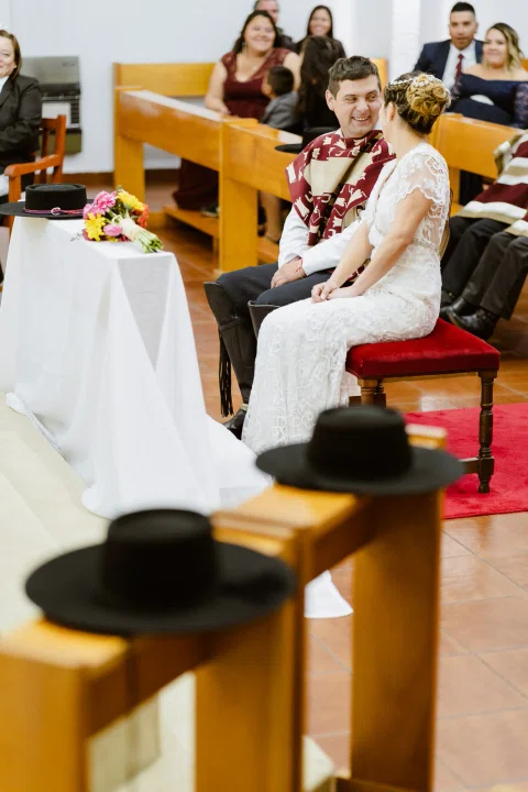 Hombre con poncho y mujer con vestido blanco están sentados juntos en un banco rojo, con sombreros tradicionales en primer plano.