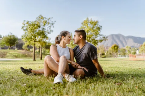 Hombre y mujer están sentados sobre el césped con árboles y montañas al fondo.