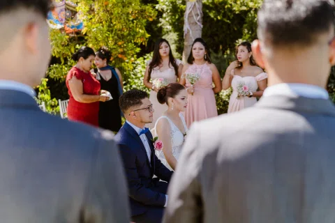 Mujer con vestido blanco y hombre con traje azul están sentados, enmarcados por dos hombres desenfocados en primer plano y varias mujeres con ramos al fondo.