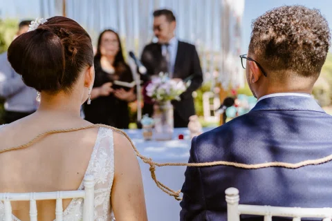 Mujer con vestido blanco y hombre con traje azul están sentados de espaldas con una cuerda rodeando sus hombros.