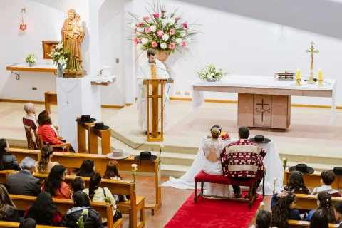 Hombre con poncho y mujer con vestido blanco están sentados frente al altar de una iglesia.