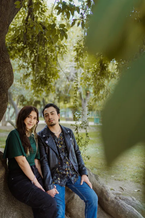Hombre y mujer están sentados juntos junto a un árbol, vistos entre hojas desenfocadas en primer plano.