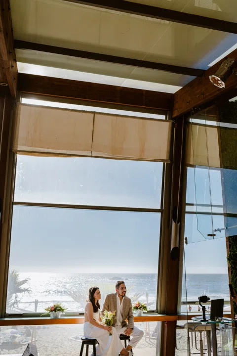 Hombre y mujer aparecen sentados a distancia junto a un gran ventanal con vista al mar.