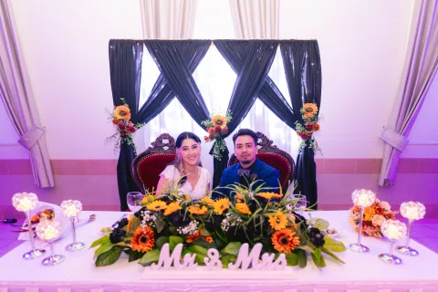 Hombre con traje azul y mujer con vestido blanco permanecen sentados detrás de una mesa decorada con flores naranjas.