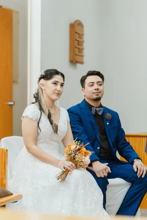 Mujer con vestido blanco y hombre con traje azul permanecen sentados y tomados de la mano en una banca.