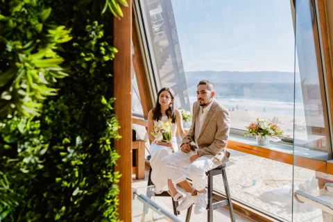 Hombre y mujer están sentados junto a un gran ventanal con vista al mar; ella sostiene un ramo.
