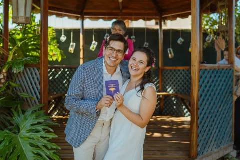 Hombre y mujer sonríen frente al gazebo mientras sostienen juntos un pequeño cuaderno morado.