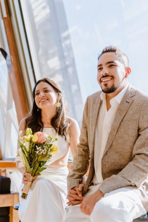 Hombre y mujer sonríen sentados frente a un ventanal; ella sostiene un ramo de flores.