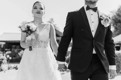 Mujer con vestido blanco y hombre con traje oscuro están tomados de la mano en una fotografía en blanco y negro; ella sostiene un ramo.