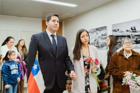 Hombre con traje oscuro y mujer de chaqueta clara se toman de la mano; ella sostiene flores rojas y una bandera de Chile aparece detrás.