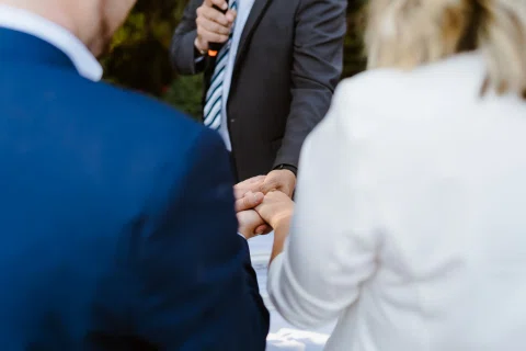 Hombre con traje azul y mujer vestida de blanco se toman de la mano frente a un hombre que sostiene un micrófono.