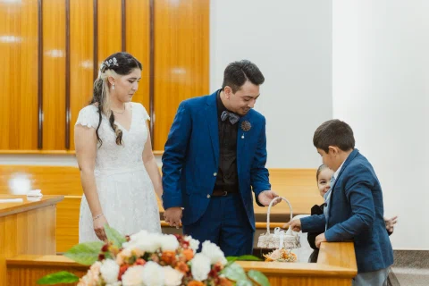Hombre con traje azul y mujer con vestido blanco se toman de la mano mientras un niño revisa una pequeña canasta y otra niña asoma detrás.