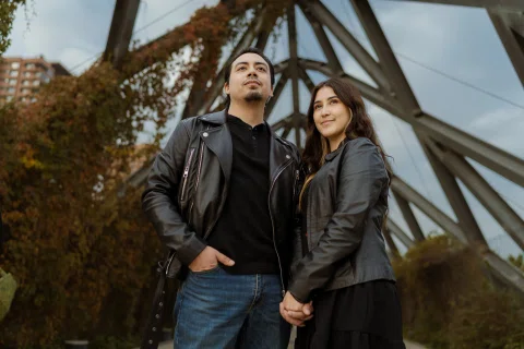 Hombre y mujer permanecen de pie tomados de la mano bajo la estructura de un puente metálico.