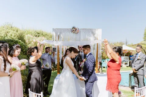 Mujer con vestido blanco y hombre con traje azul se toman de las manos mientras dos mujeres sostienen una tela blanca extendida sobre ellos.