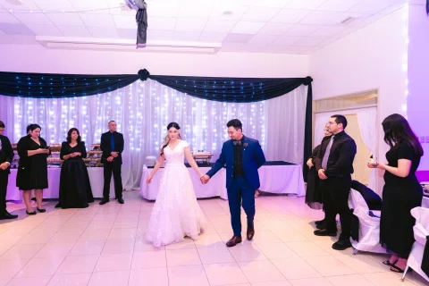 Hombre con traje azul y mujer con vestido blanco se toman de las manos en el centro de un salón, rodeados por varias personas.