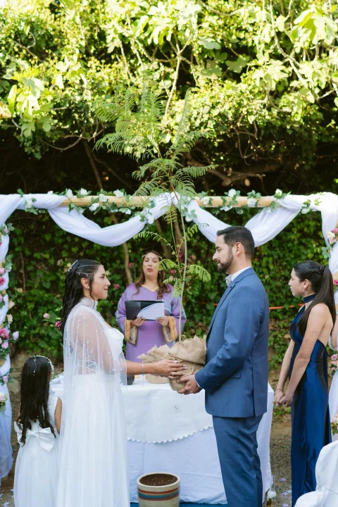 Hombre con traje azul y mujer con vestido blanco se toman de las manos frente a una mujer con vestido morado bajo un arco floral.