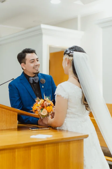 Hombre con traje azul y mujer con vestido blanco se miran tomados de las manos junto a un atril.