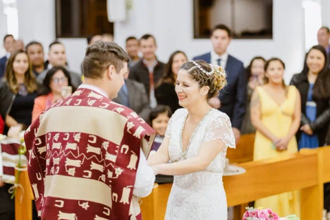 Hombre con poncho y mujer con vestido blanco se toman de las manos dentro de una iglesia, rodeados por otras personas.